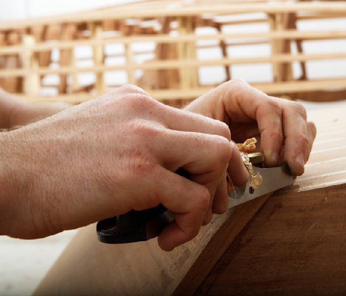 Hands working on a wooden guitar neck with a file tool.