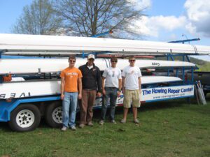 Four men standing in front of a rowing boat on a grassy area.