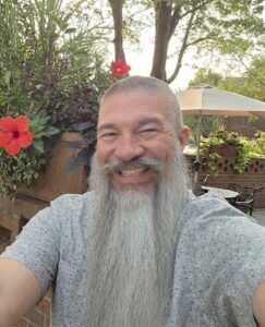 Smiling man with a long grey beard sitting outdoors near flowers.