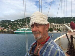 Man in a white hat smiles near a harbor with boats and hills in the background.