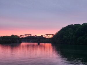 A serene sunset over a river with a silhouetted bridge and trees.