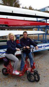 Two people smiling on a tandem bike outside a boat repair center.