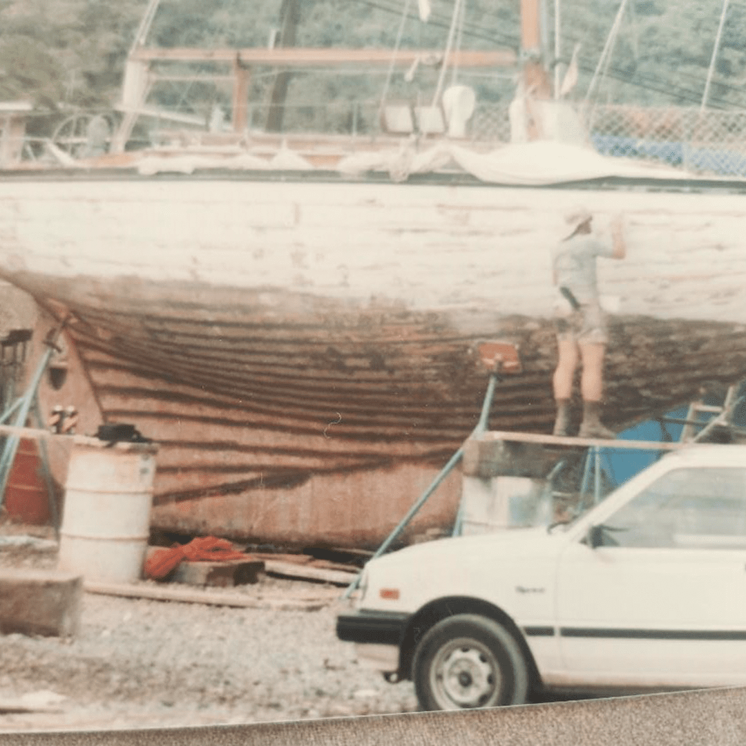 A man works on a large wooden boat beside a white car in a yard.