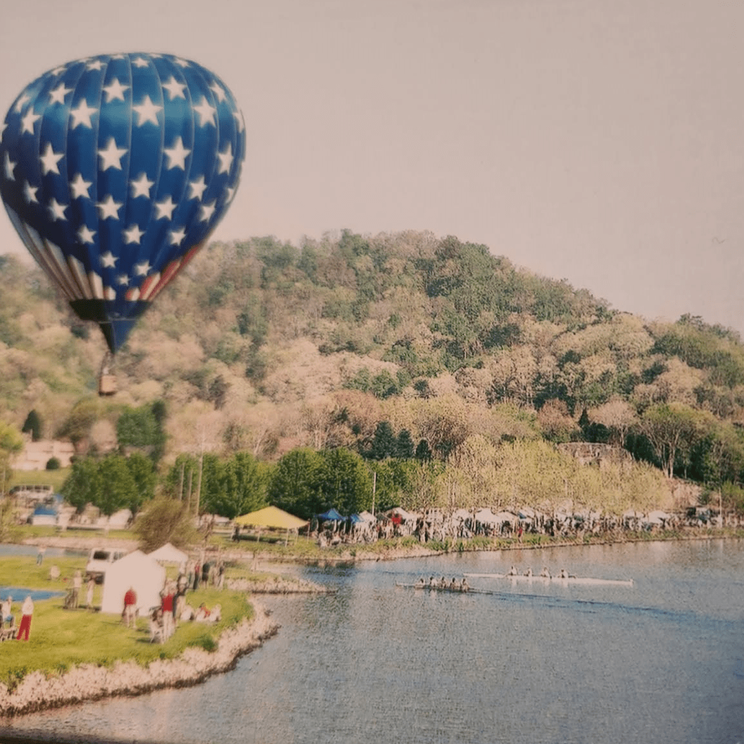 Hot air balloon flying over a lakeside park with hills in the background.