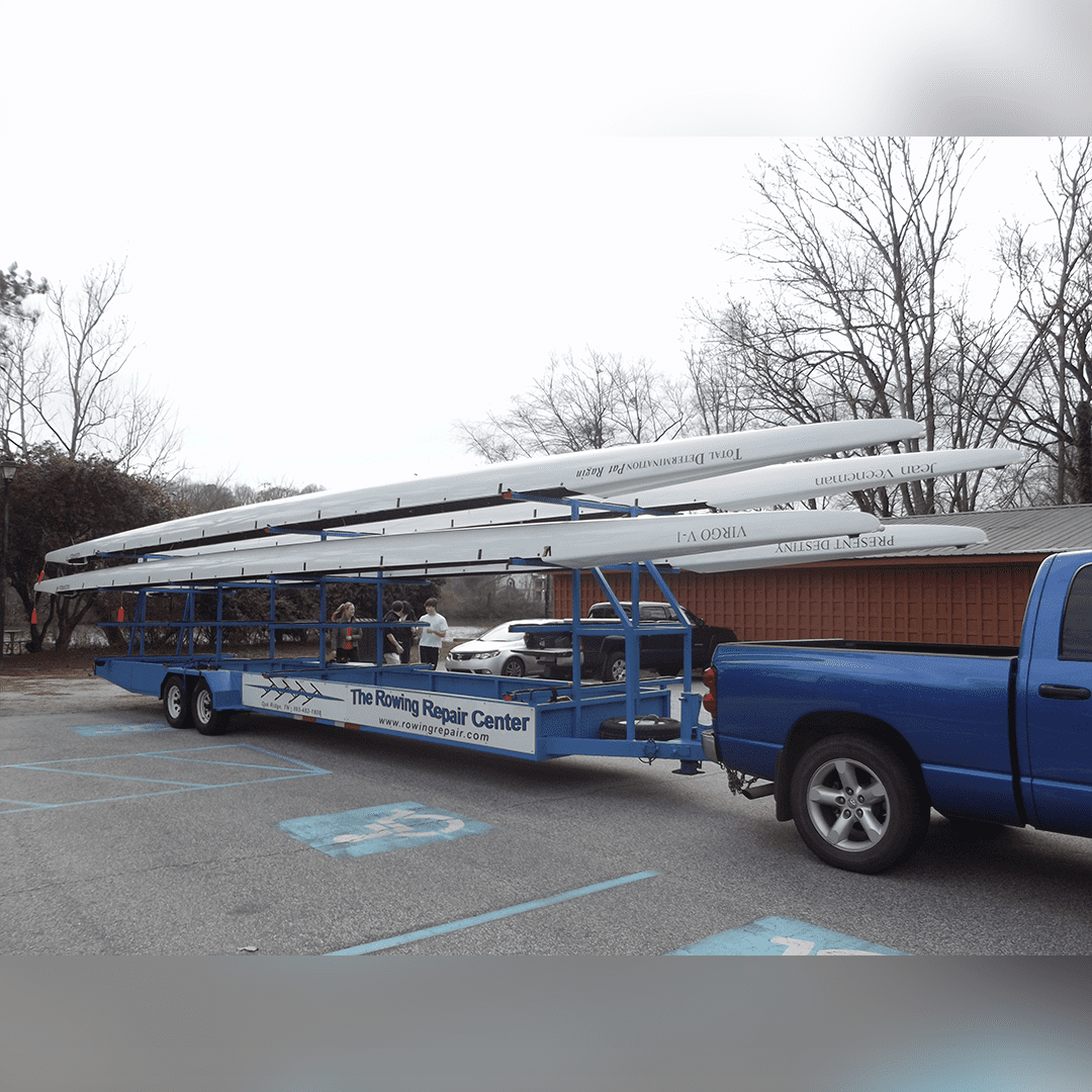 Blue pickup truck towing a trailer loaded with long poles in a parking lot.