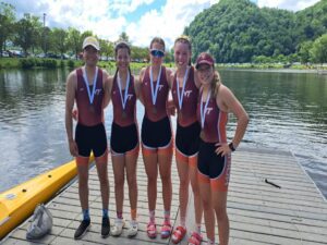 Five female rowers posing with medals by a lake and mountains.